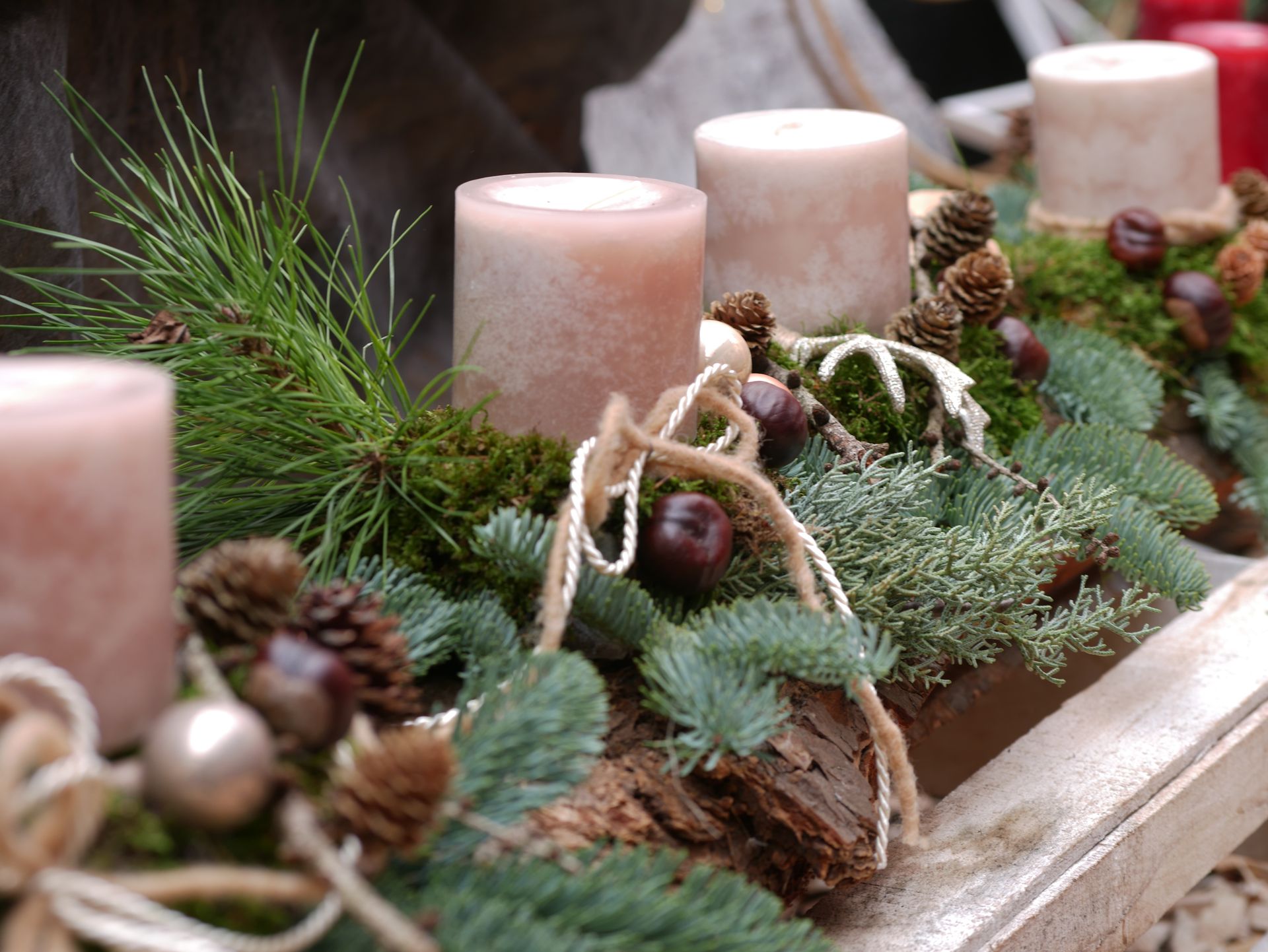 Mehrere beige Kerzen, umgeben von grünem Tannengrün, Kiefernzweigen, Tannenzapfen und dekorativen Naturmaterialien, angeordnet auf einem Holzbalken als Adventskranz.