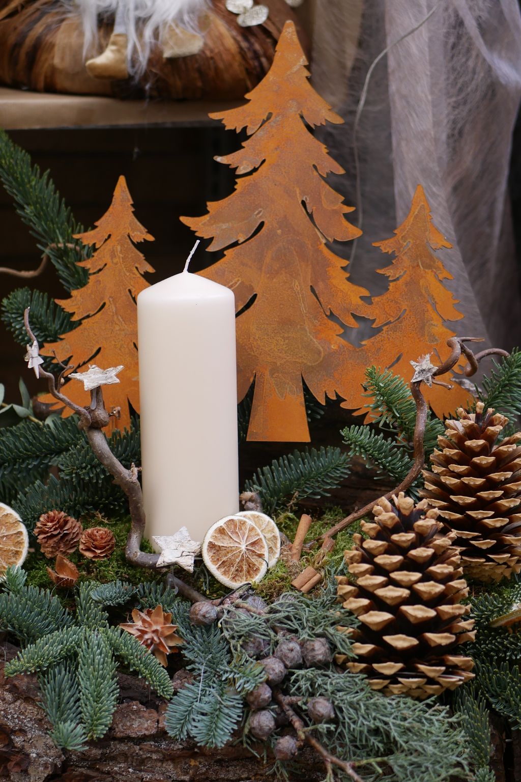 Weihnachtliche Dekoration mit einer weißen Kerze, getrockneten Orangenscheiben, Tannenzapfen und dekorativen Metalltannenbäumen aus Roststahl, umgeben von grünen Tannenzweigen.