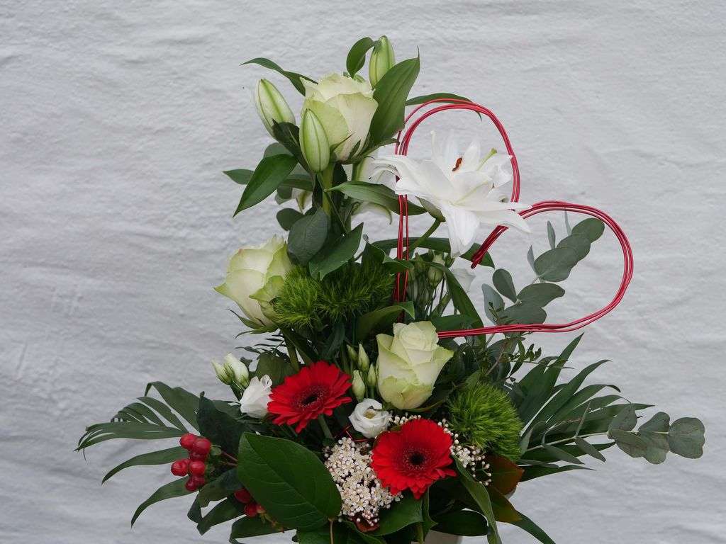Ein buntes Blumenarrangement mit weißen Rosen, weißen Lilien, roten Gerbera und grünen Blättern vor einem weißen Hintergrund, dekoriert mit einem roten, herzförmigen Draht.