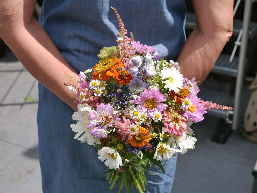 Person im blauen Kleid hält einen bunten Blumenstrauß aus verschiedenen Blumenarten vor dem Körper.