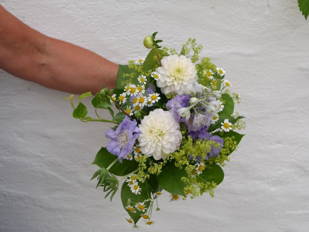 Eine Hand hält einen kleinen Blumenstrauß mit weißen Dahlien, lila und gelben Blüten sowie grünen Blättern vor einer weißen Wand.