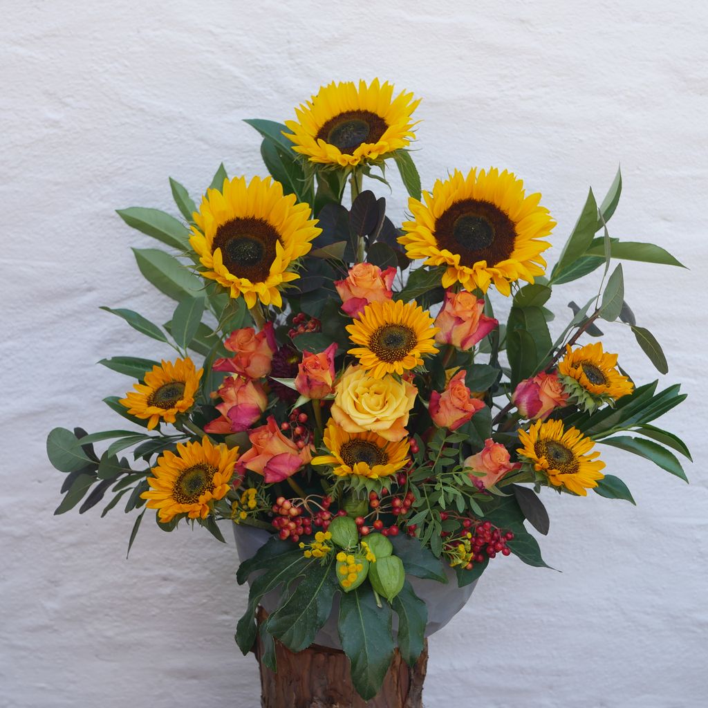 Bunter Blumenstrauß mit gelben Sonnenblumen, gelben und orangefarbenen Rosen sowie grünen Blättern vor einer weißen Wand.