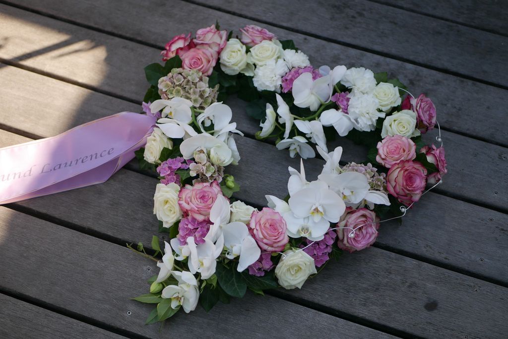 Herzförmiger Blumenkranz aus weißen Orchideen, rosa und weißen Rosen sowie anderen Blüten, auf einer grauen Holzdiele liegend, mit einem rosafarbenen Schleifenband.