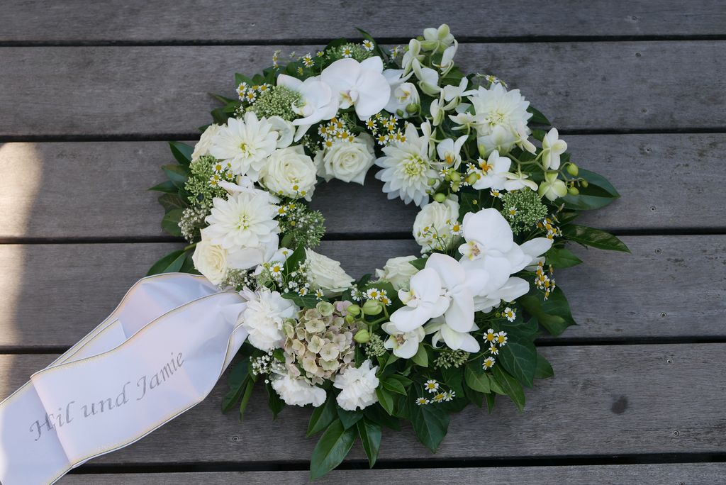 Kranz aus weißen Blumen mit grünen Blättern und einer weißen Schleife mit der Aufschrift auf einer Holzoberfläche.