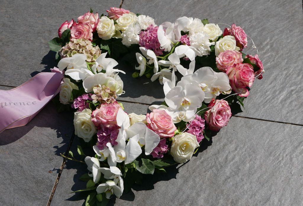 Herzförmiges Blumenarrangement mit weißen Orchideen, rosa und weißen Rosen sowie lila Hortensien auf einem grauen Steinboden.