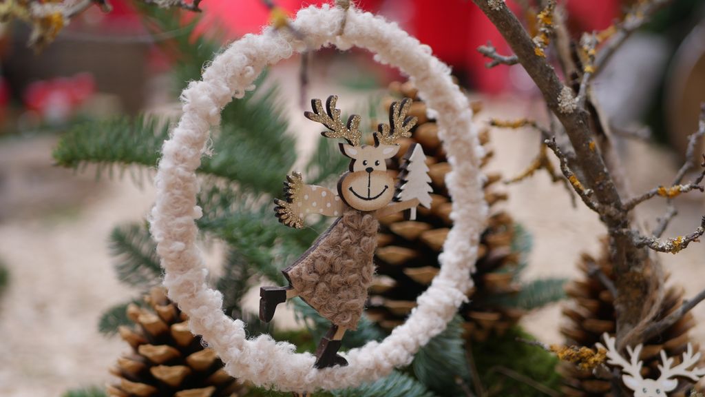 Ein hängender handgemachter Rentieranhänger mit einem weißen Tannenbaum, umrahmt von einem weißen, wolligen Ring vor einem Hintergrund aus Tannenzapfen und Tannenzweigen.