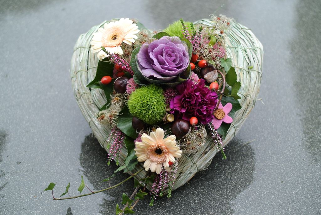 Herzförmige Blumenarrangement mit verschiedenen Blumen wie lila Zierkohl, rosa Nelken, cremefarbenen Gerbera und grünen Kugelmoos auf grauem Untergrund.