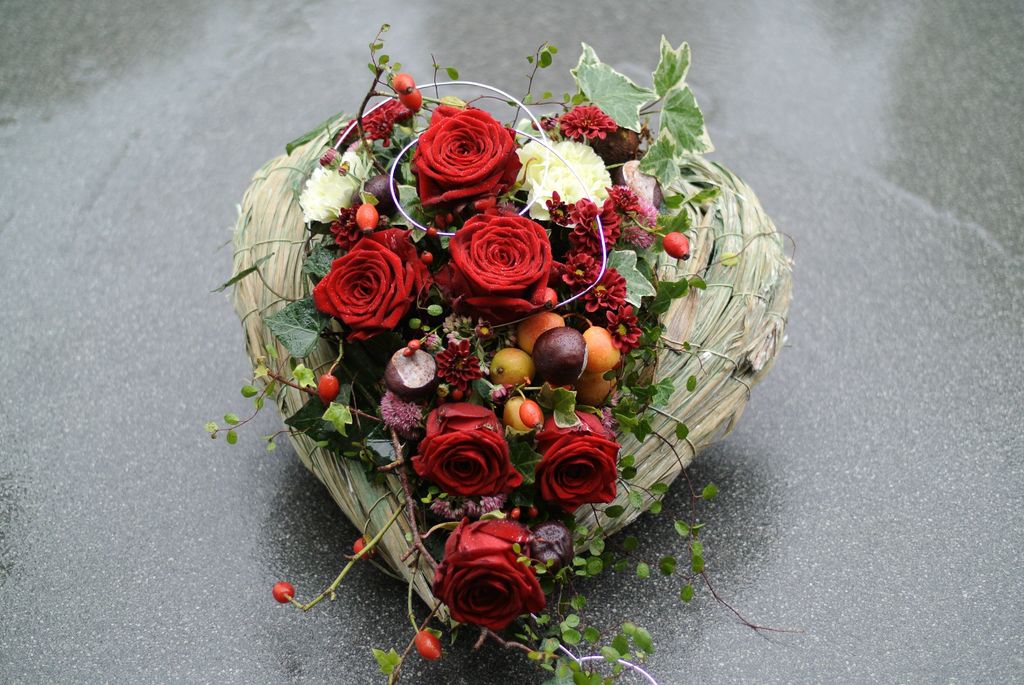 Ein herzförmiges Blumenarrangement aus roten Rosen, weißen und roten Blumen, Beeren und grünen Blättern auf einem dunklen Untergrund.