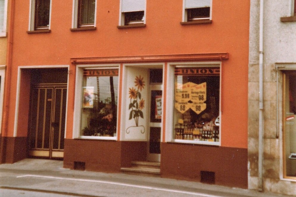 Außenansicht eines kleinen Ladengeschäfts mit orangefarbener Fassade, zwei Schaufenstern und einem Eingang in der Mitte, mit Sonnblumenwandmalerei am Türrahmen.