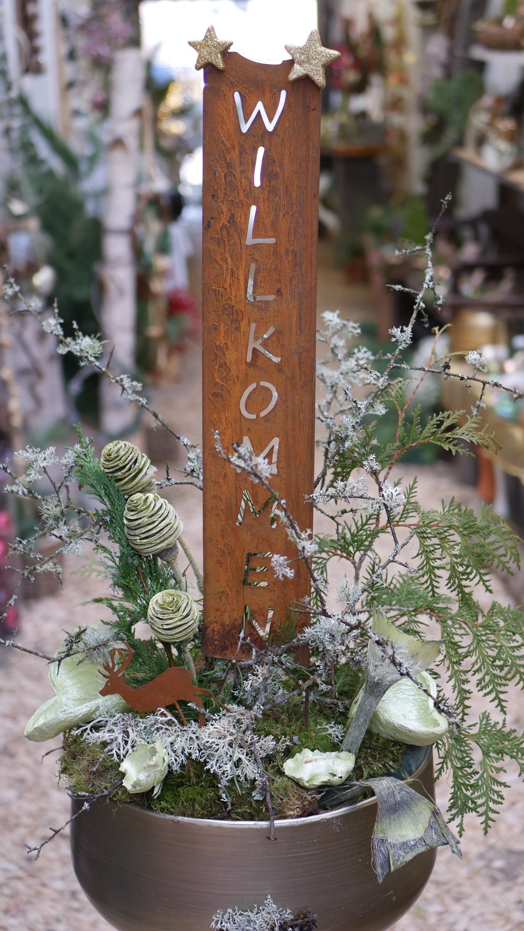 Dekorativer Pflanzgefäß mit einem rostigen Holzschild darauf, auf dem senkrecht das Wort 'WILLKOMMEN' steht, verziert mit trockenen Blumen, Moos und einem kleinen Hirsch aus Metall.