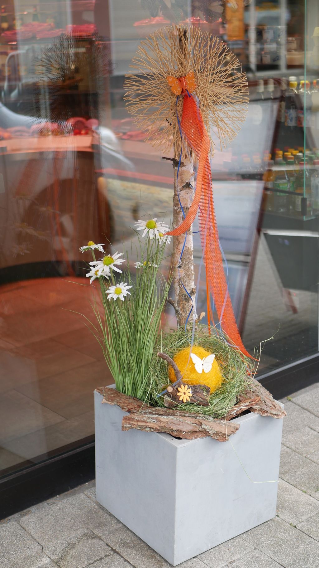 Blumendekoration in einem grauen quadratischen Pflanzkasten vor einem Geschäft, bestehend aus Gras, weißen Gänseblümchen, einem Birkenstamm mit orangefarbenem Netzband und einem gelben Ball mit weißem Schmetterling.