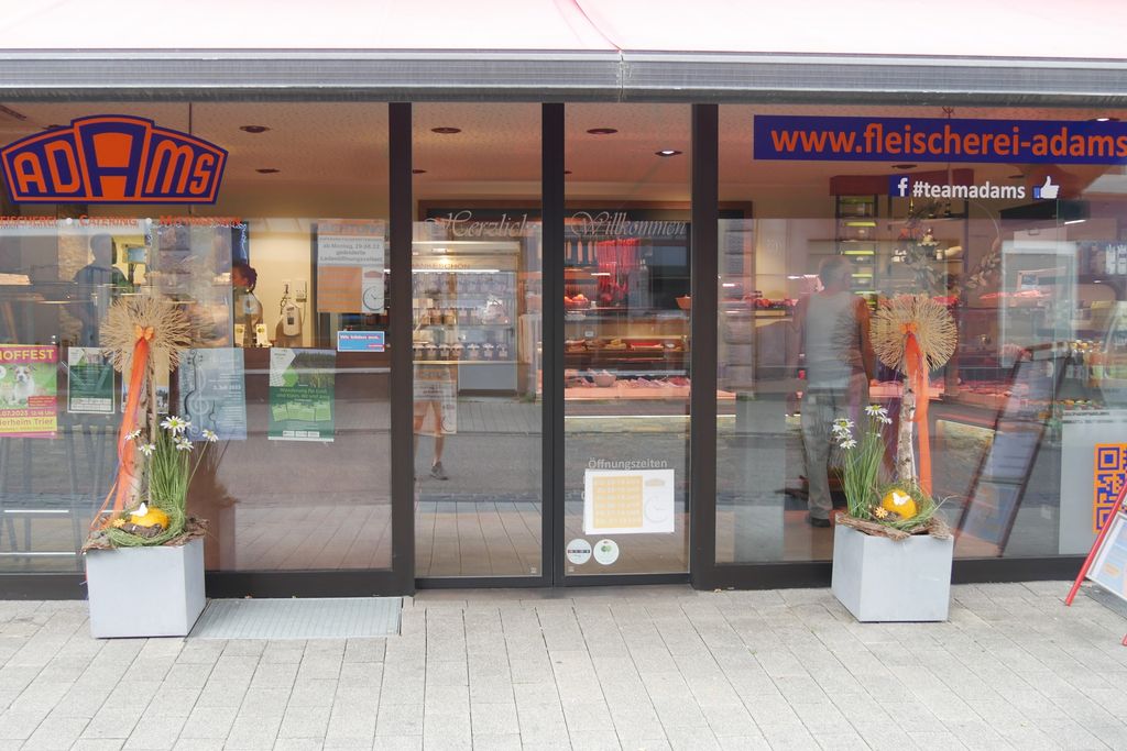 Eingangsbereich einer Fleischerei mit gläserner Schiebetür, Dekoration mit Blumen und Kürbissen links und rechts, dahinter Sicht auf Auslage mit Fleischwaren und eine Person im Geschäft.