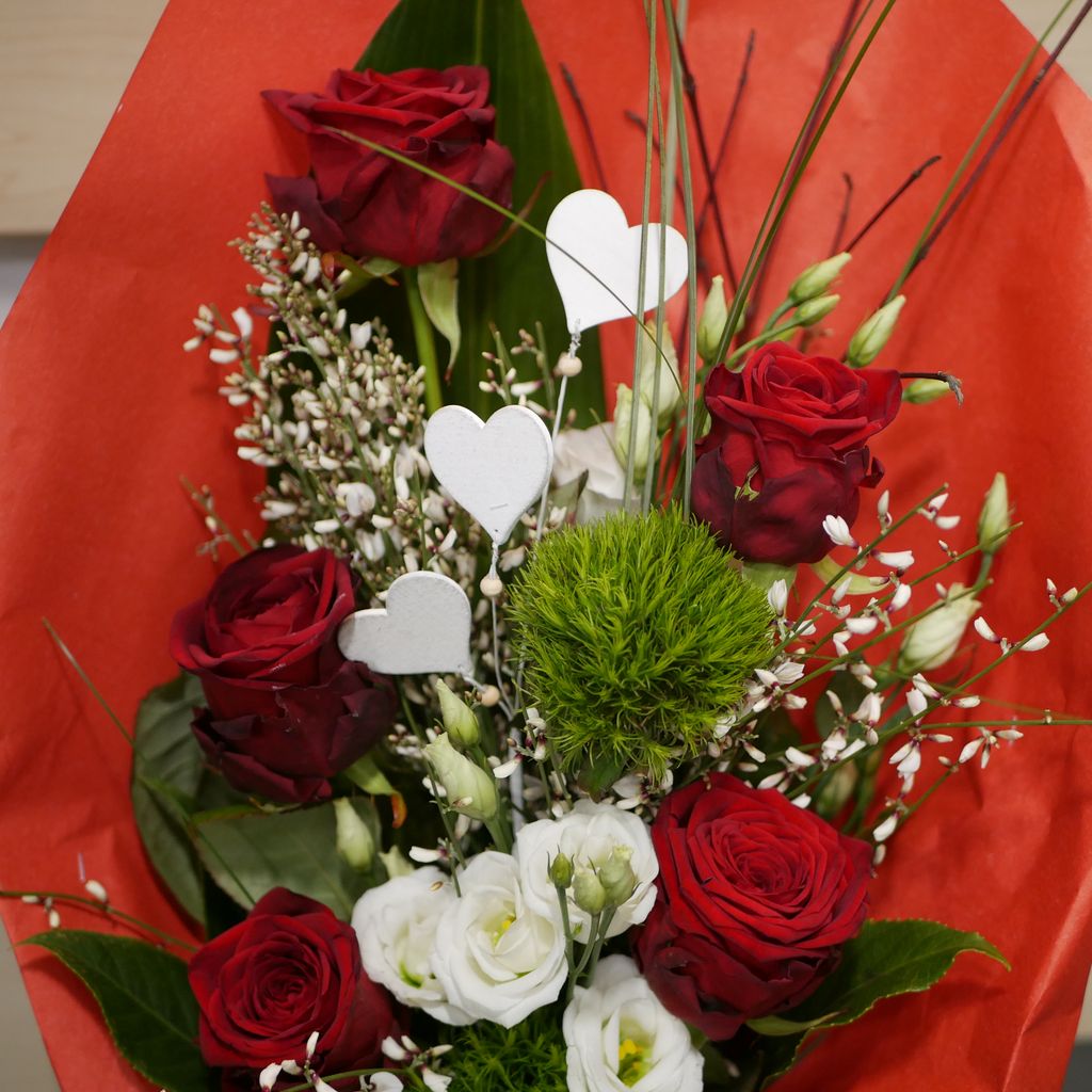 Strauß aus roten Rosen, weißen Blumen und grünen Blättern, dekoriert mit weißen Herzfiguren, vor einem roten Hintergrund.